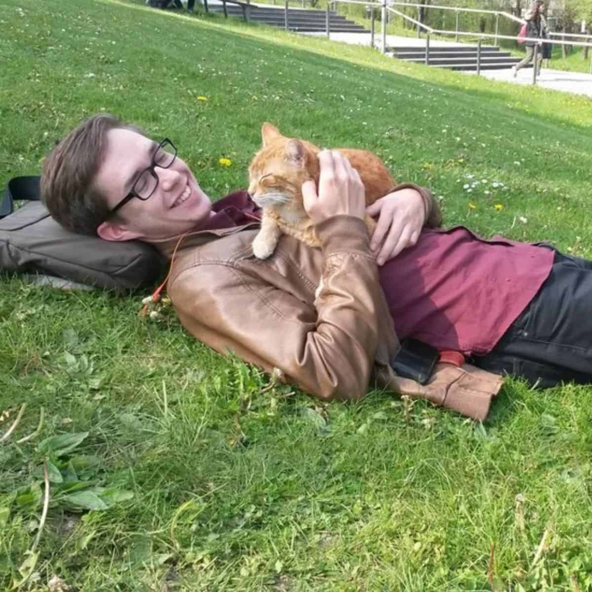 a smiling man is lying on the grass and petting a cat