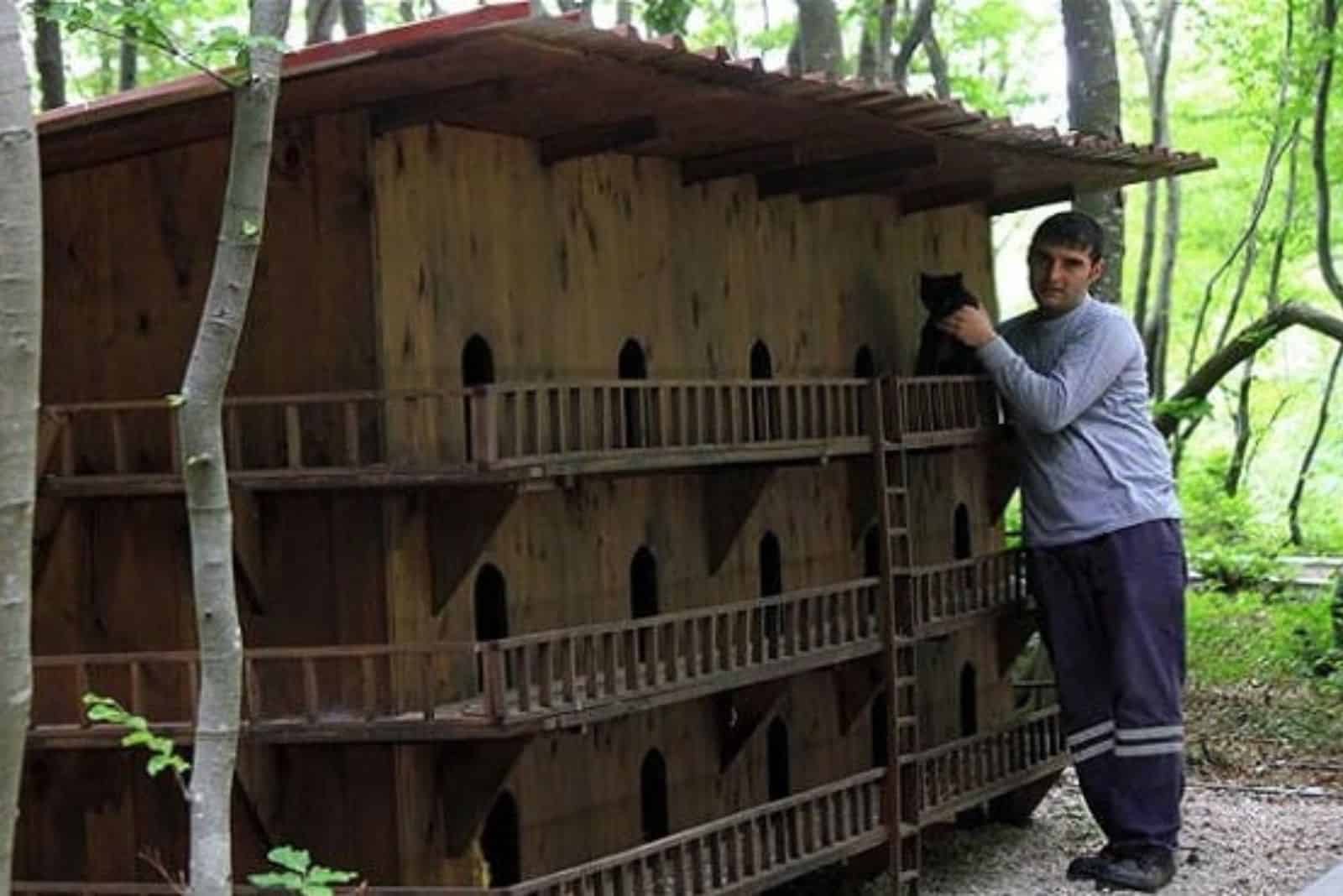 photo of a man holding a cat next to a cat house