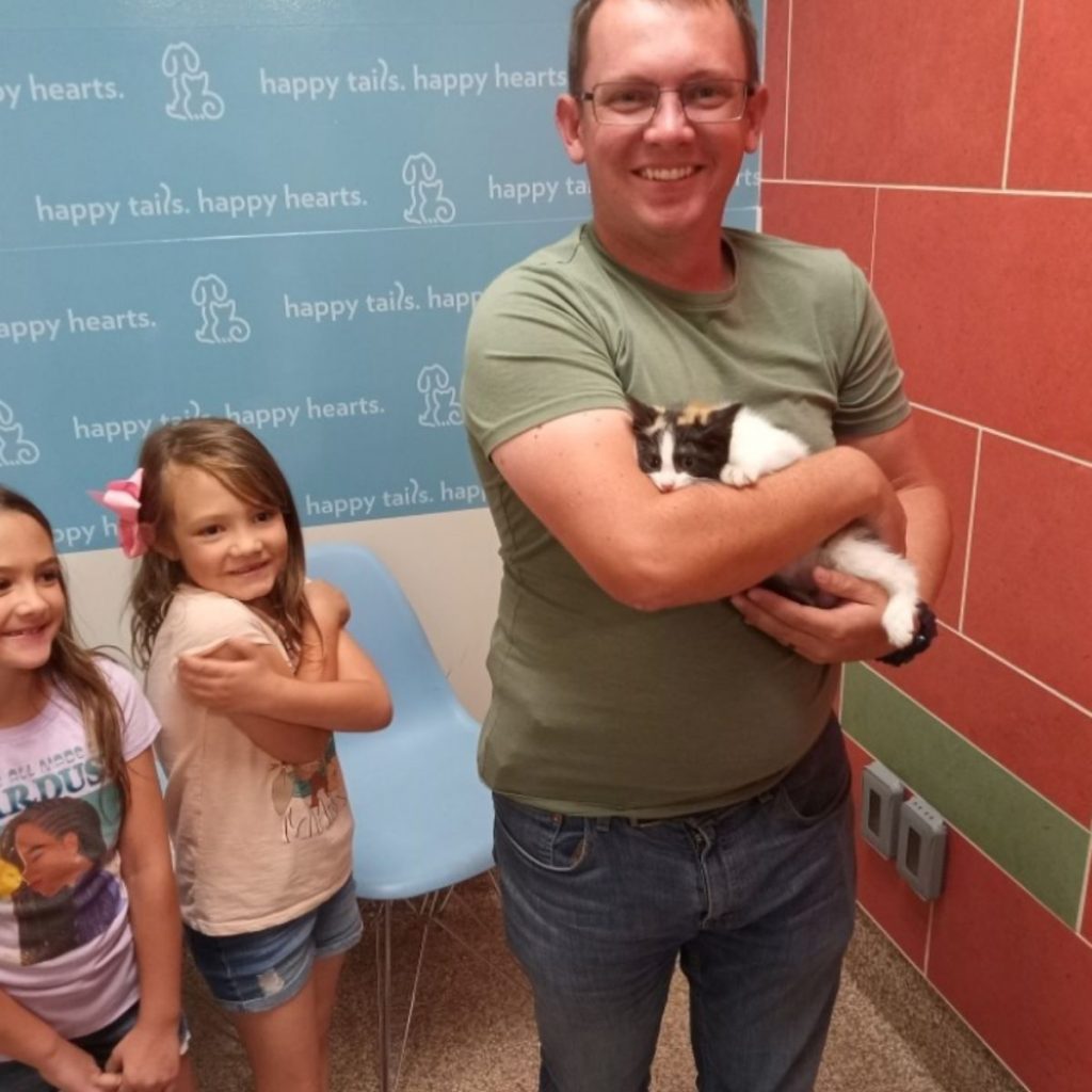 two cute little girls are standing next to their father who is holding a kitten in his arms