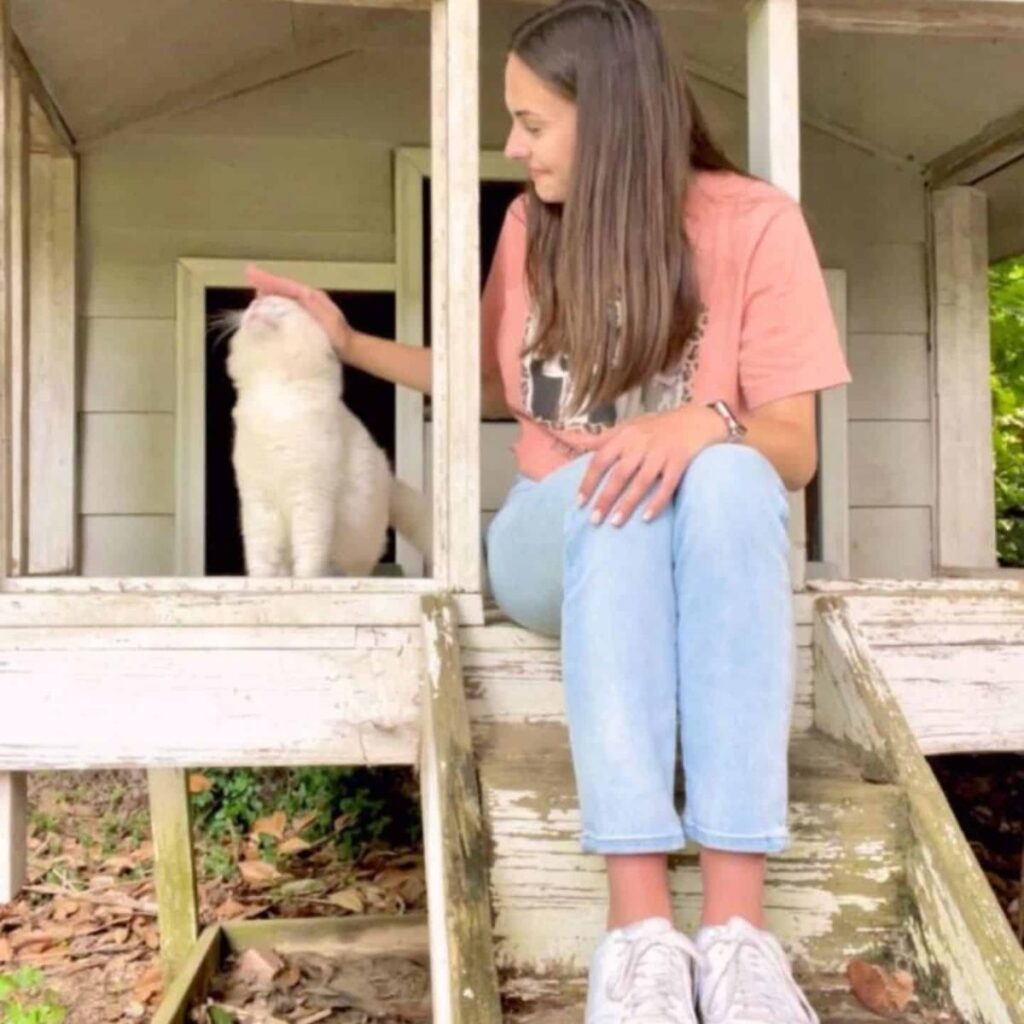 A girl sits next to a cat in front of a house