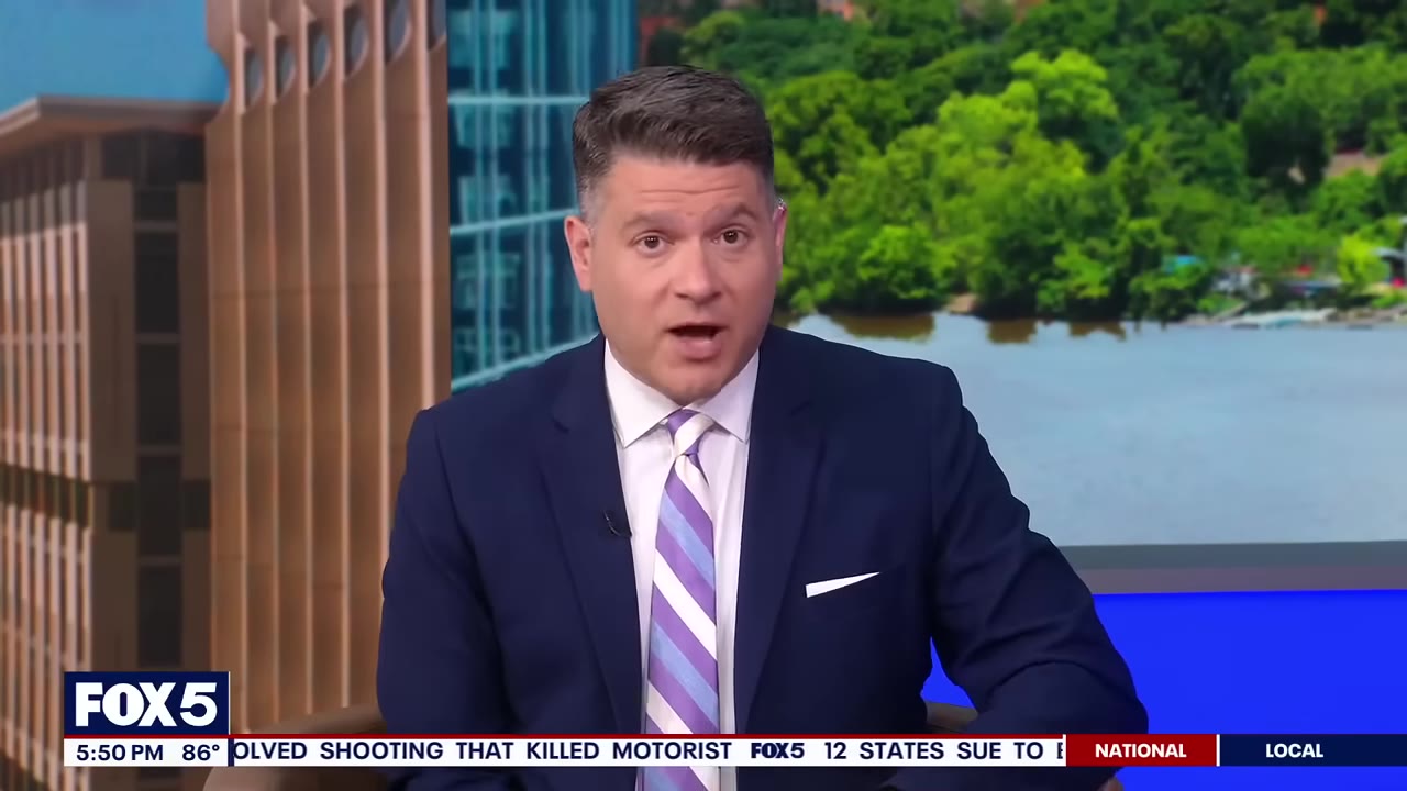 A news anchor in a blue suit and striped tie speaks with a serious expression in front of a studio background showing a river and trees.