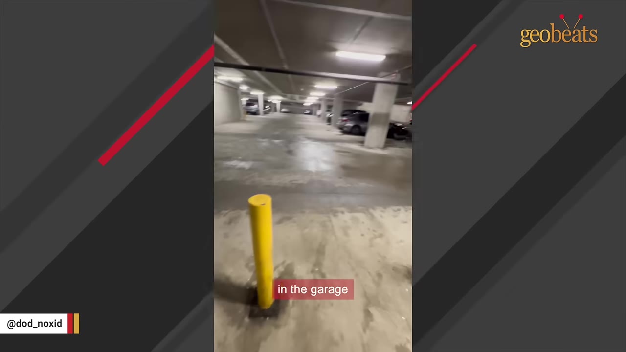 A wide shot of an indoor concrete parking garage with parked cars and overhead fluorescent lights.