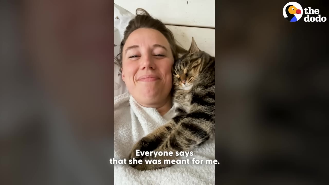 A smiling woman lies down with a tabby cat snuggled closely against her cheek.