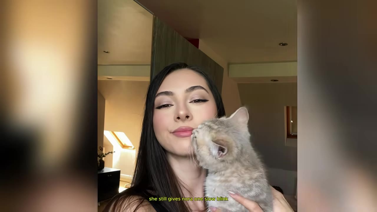A young woman with long dark hair smiling gently as a small, light-colored kitten nuzzles her cheek.
