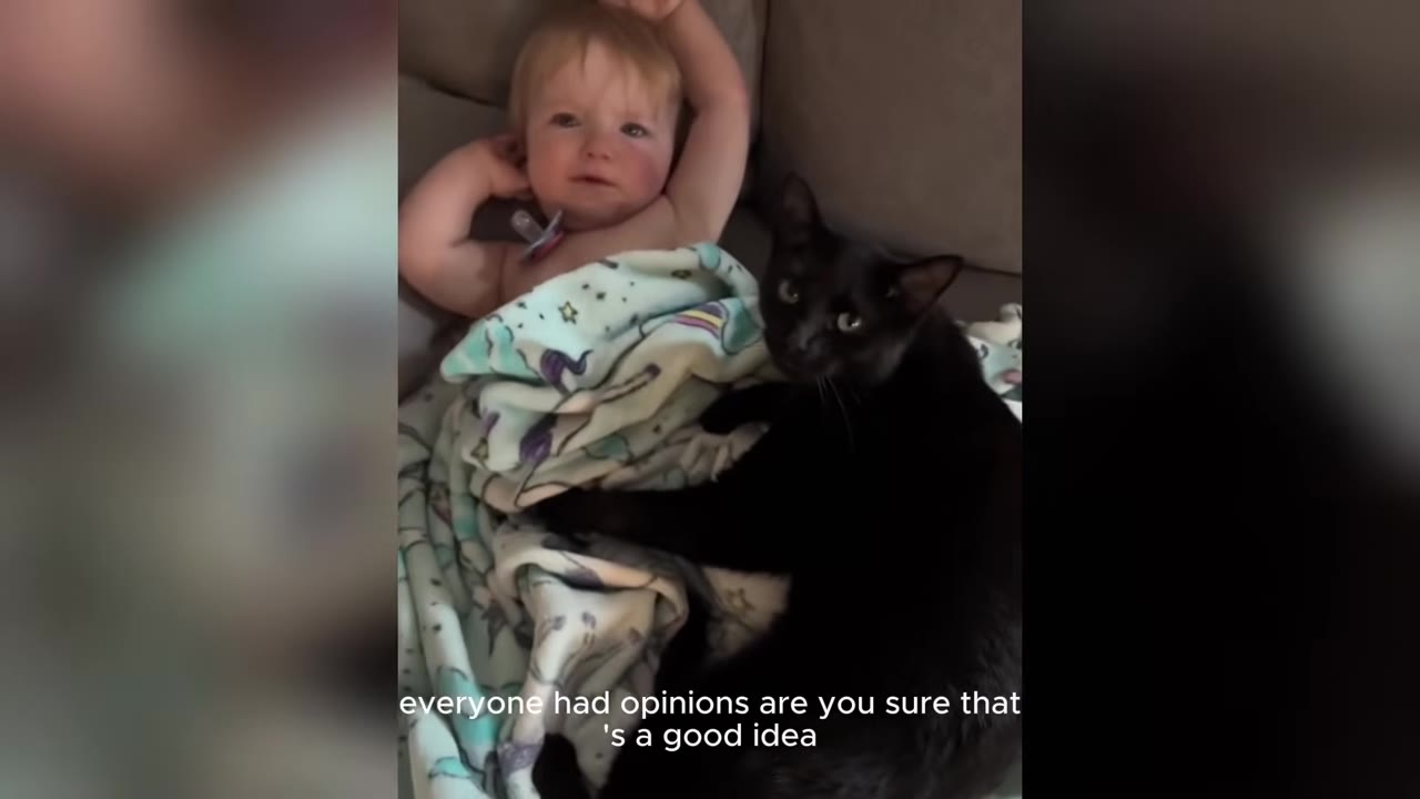 A black cat with bright, steady eyes lies on a couch next to a baby under a blanket, looking directly at the camera.