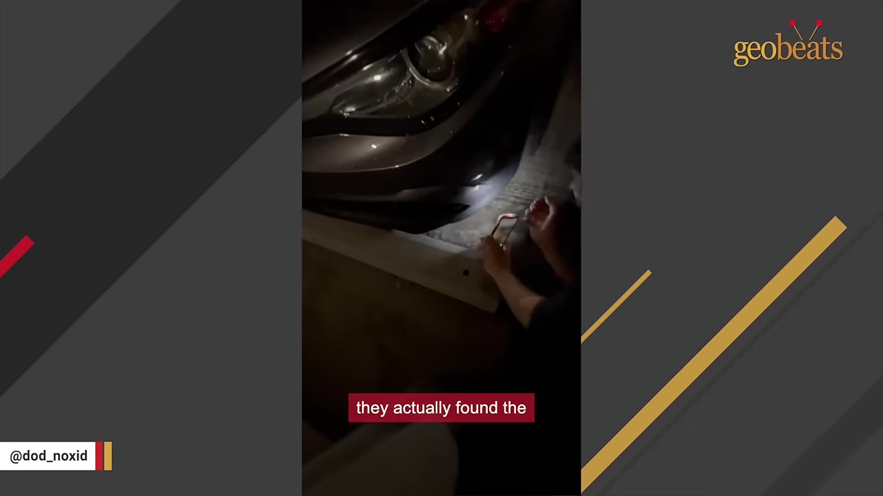 A person kneeling next to the front bumper of a car at night, using a smartphone flashlight to look underneath the vehicle.