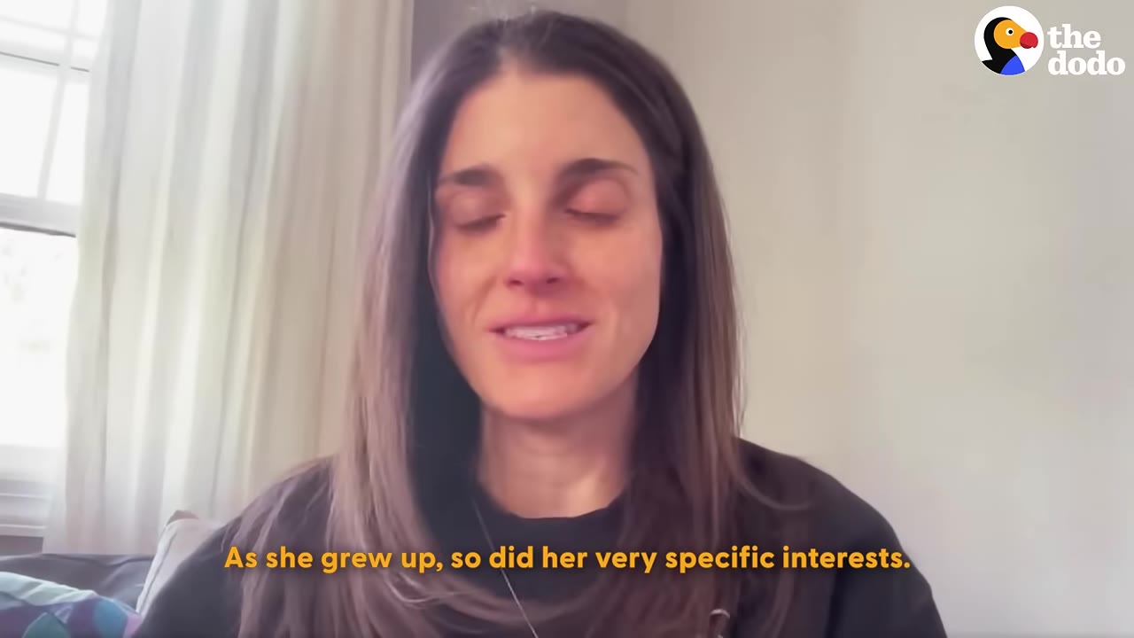 A woman with long brown hair looks directly at the camera while speaking, with a window and curtains in the background.