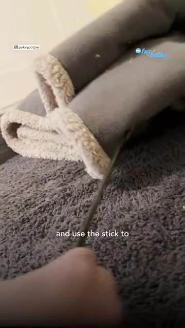 A hand holding a thin stick and pointing it toward a folded grey blanket with a white fleece lining on a grey carpet.