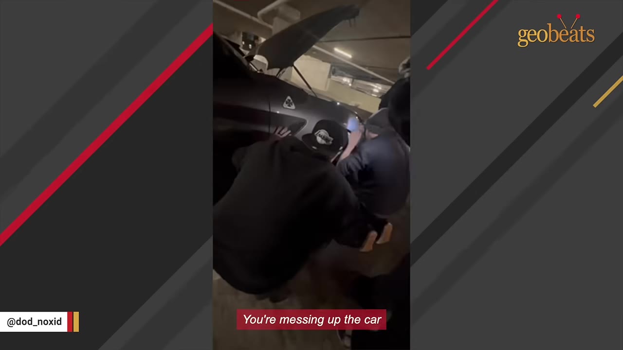 Two men crouched next to the front wheel of a dark car with its hood open in a parking garage, working to access the vehicle.