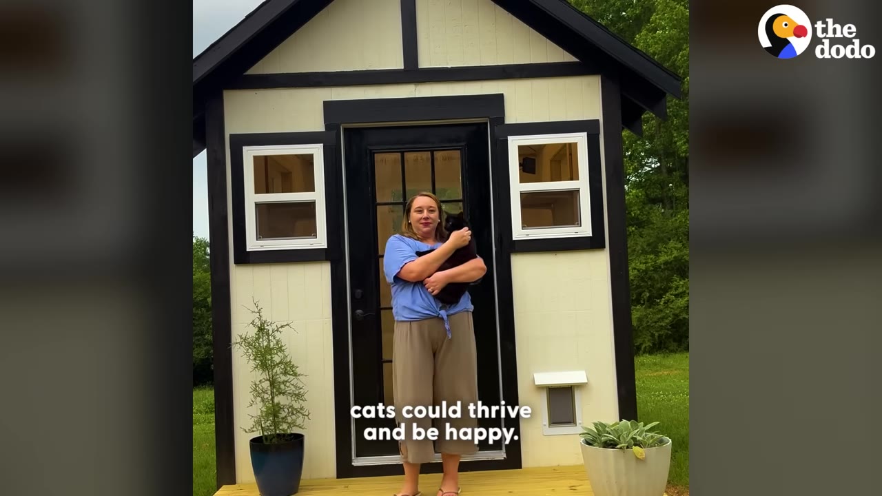 A woman stands on the porch of a small, custom-built cat house with two front windows and a glass door, holding a black cat in her arms.