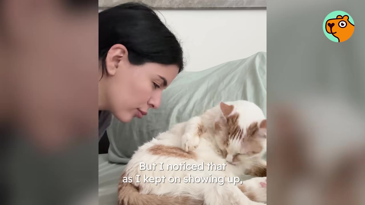 A woman with dark hair looks gently at a white and tan cat lying on a bed.