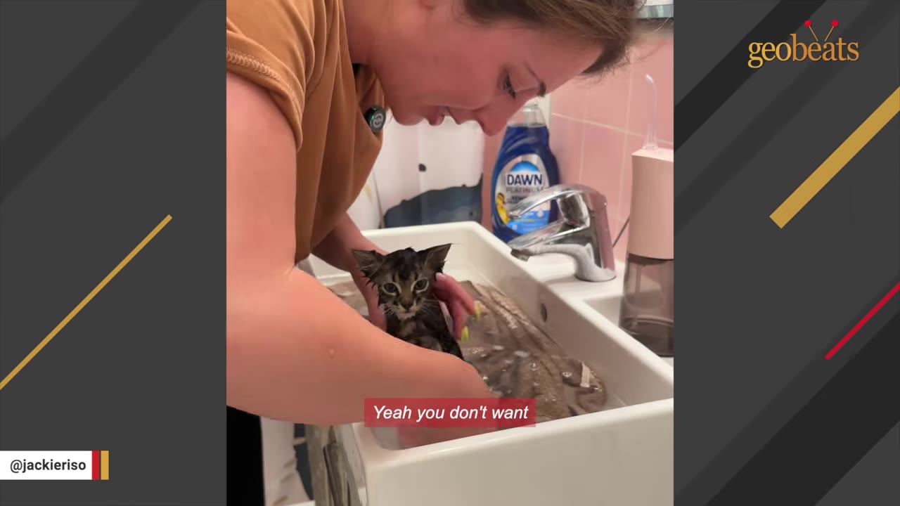 A woman gently bathes a small, wet tabby kitten in a bathroom sink while looking down at it.