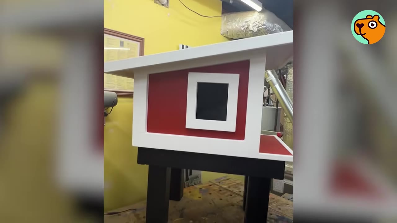 A custom-built miniature red and white wooden house with a slanted roof, sitting on black legs in a workshop.