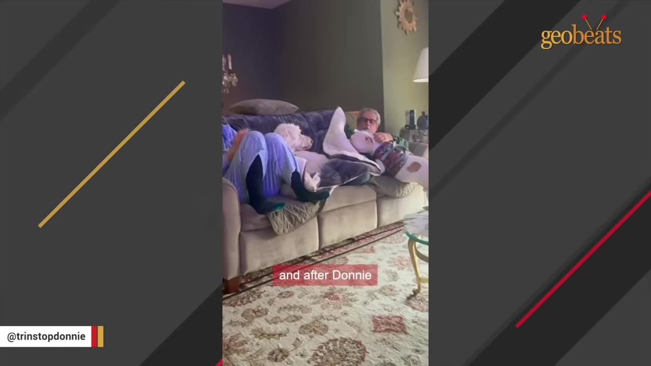 A white dog wearing a sweater cuddles on a living room couch with a man and another person.