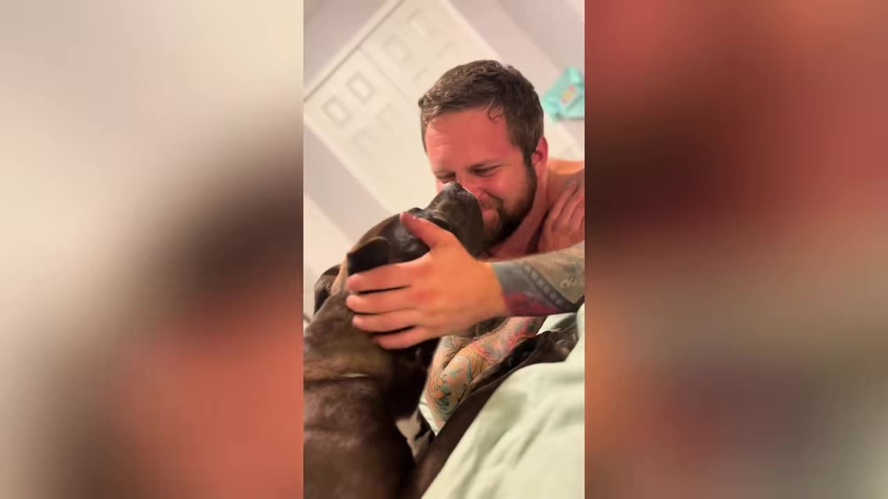 A smiling man with tattoos on his arm gently cradles a dark brindle dog's head as they share a close, affectionate moment.