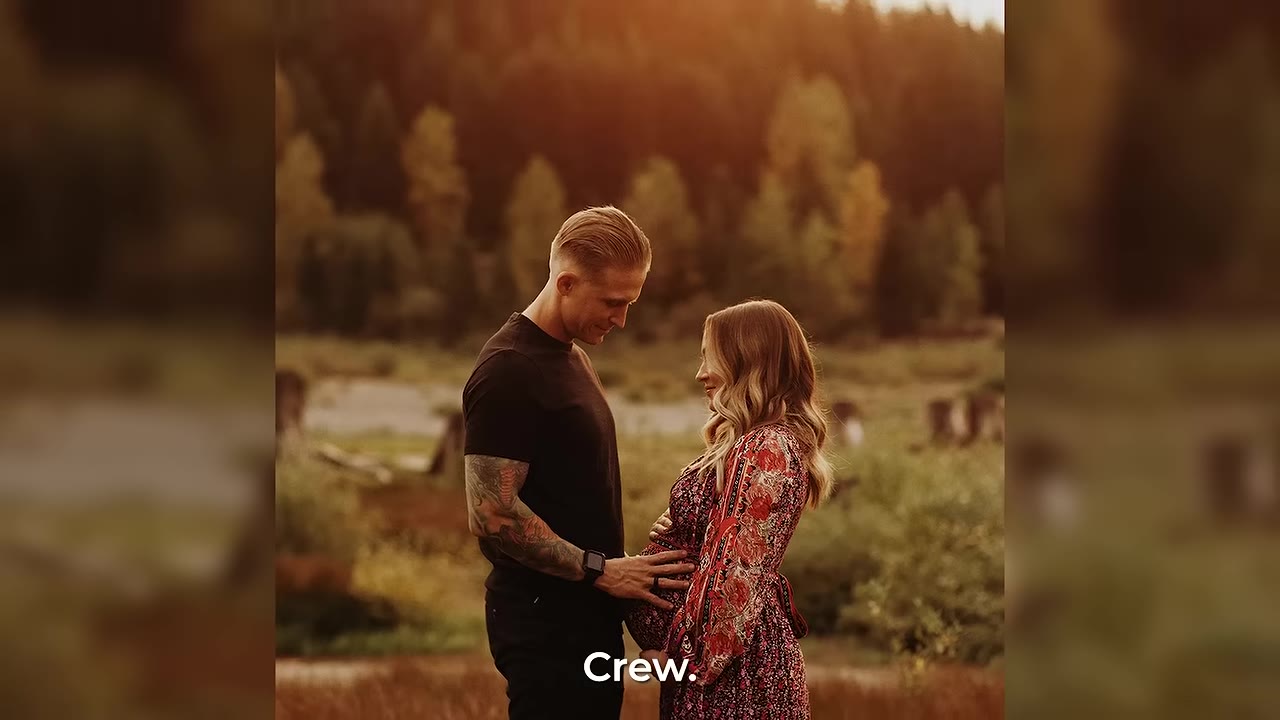 A man with tattoos on his arm gently places his hand on a pregnant woman's belly as they stand facing each other in a warm, sunlit outdoor field.