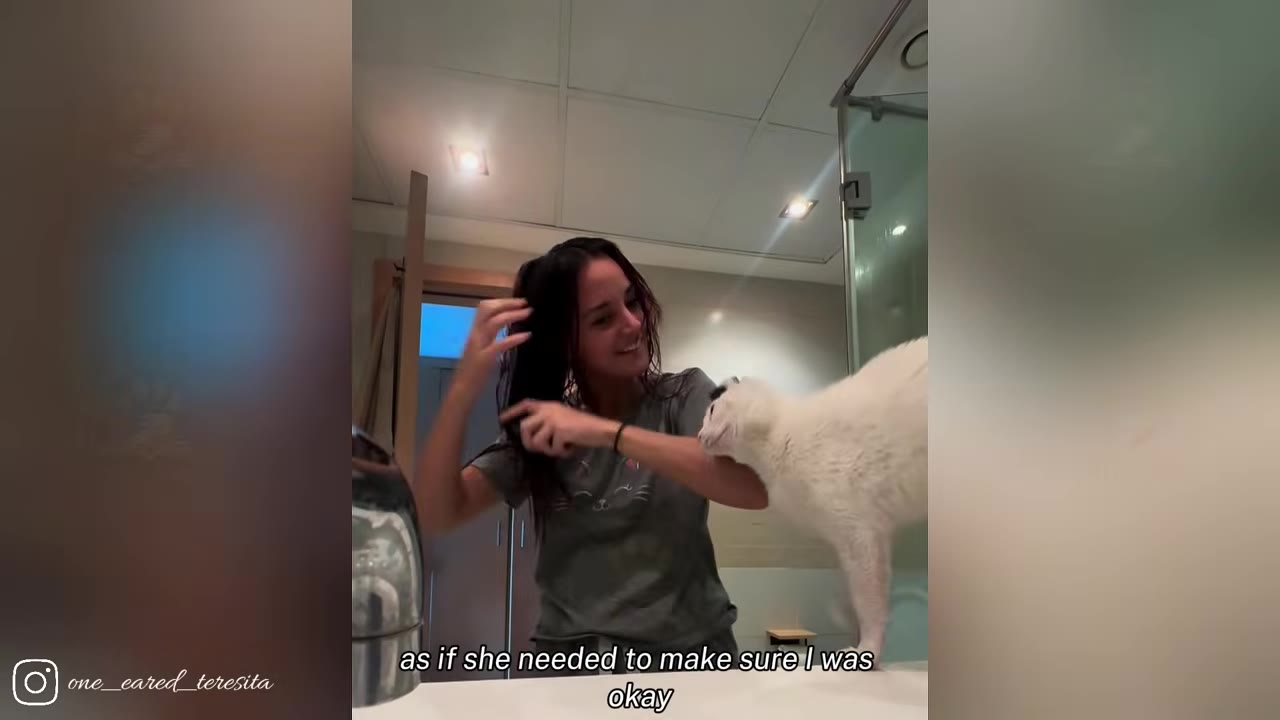 A woman with dark hair smiles as she brushes her hair in a bathroom, while a white cat stands on the counter next to her, leaning against her arm.
