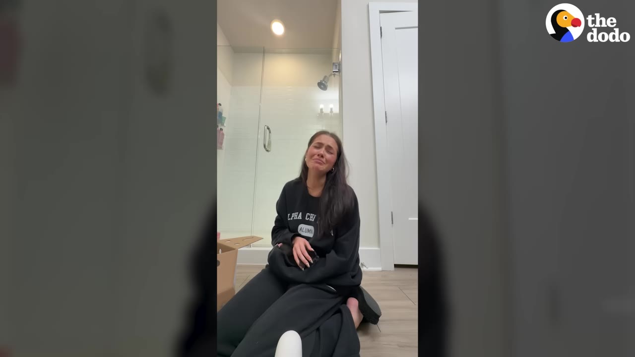 A woman sits on a bathroom floor wearing a black sweatshirt, holding a small black kitten in her lap with an emotional expression.