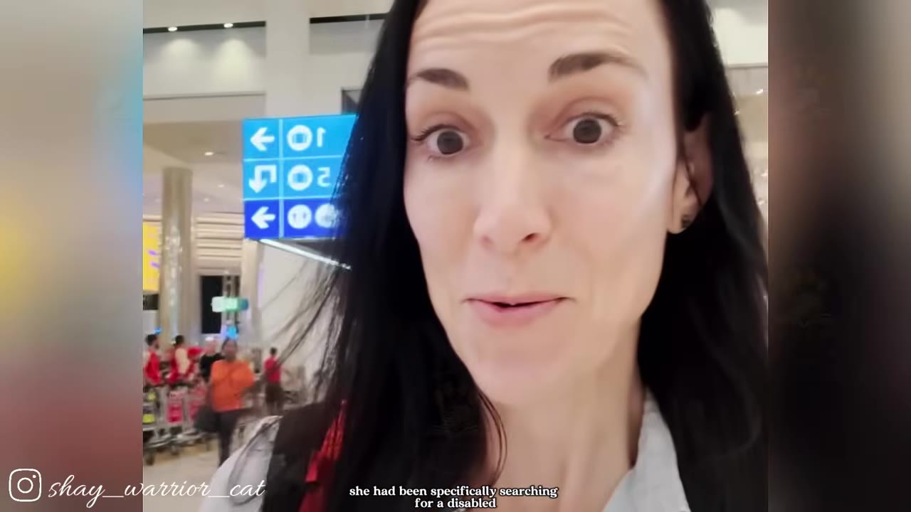 A close-up selfie video frame of a woman with long dark hair looking directly at the camera inside what appears to be an airport terminal.