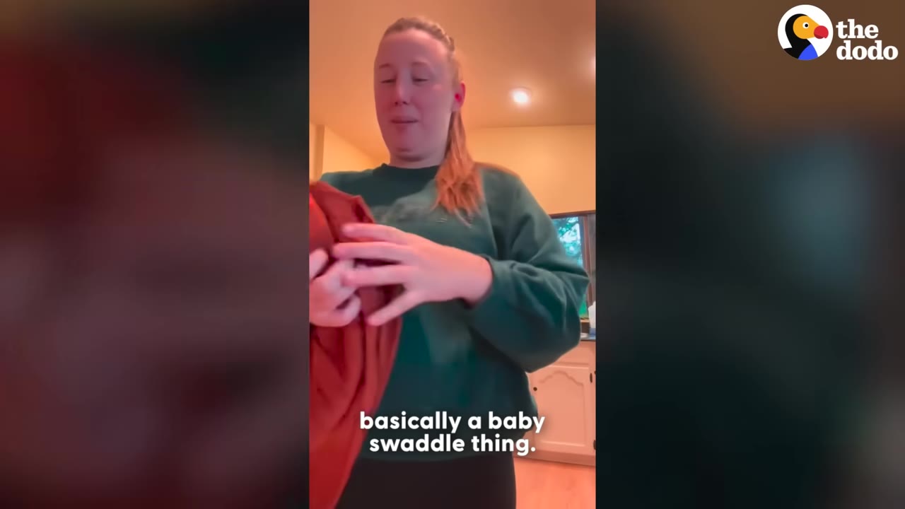 A woman in a green sweatshirt stands in a kitchen holding up an orange fabric baby swaddle wrap.