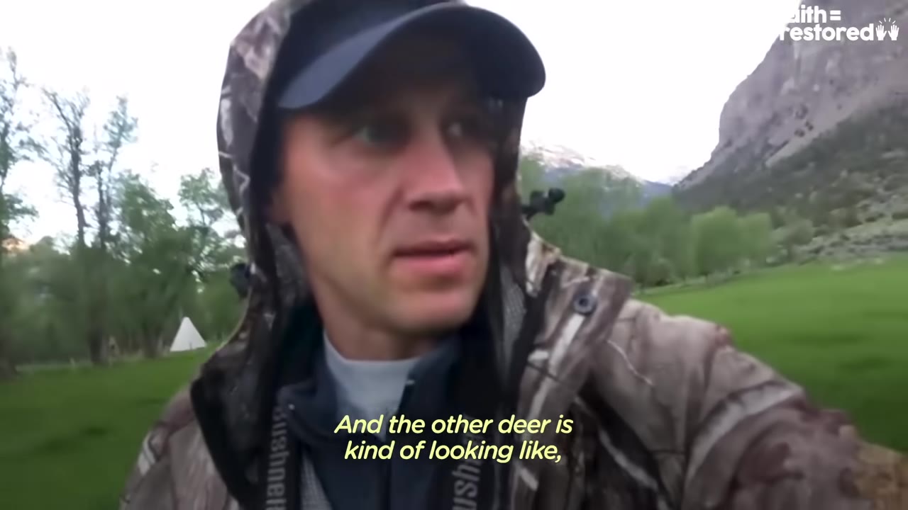 A man wearing a camouflage jacket and a baseball cap looks off-camera while standing in a green meadow with mountains in the background.