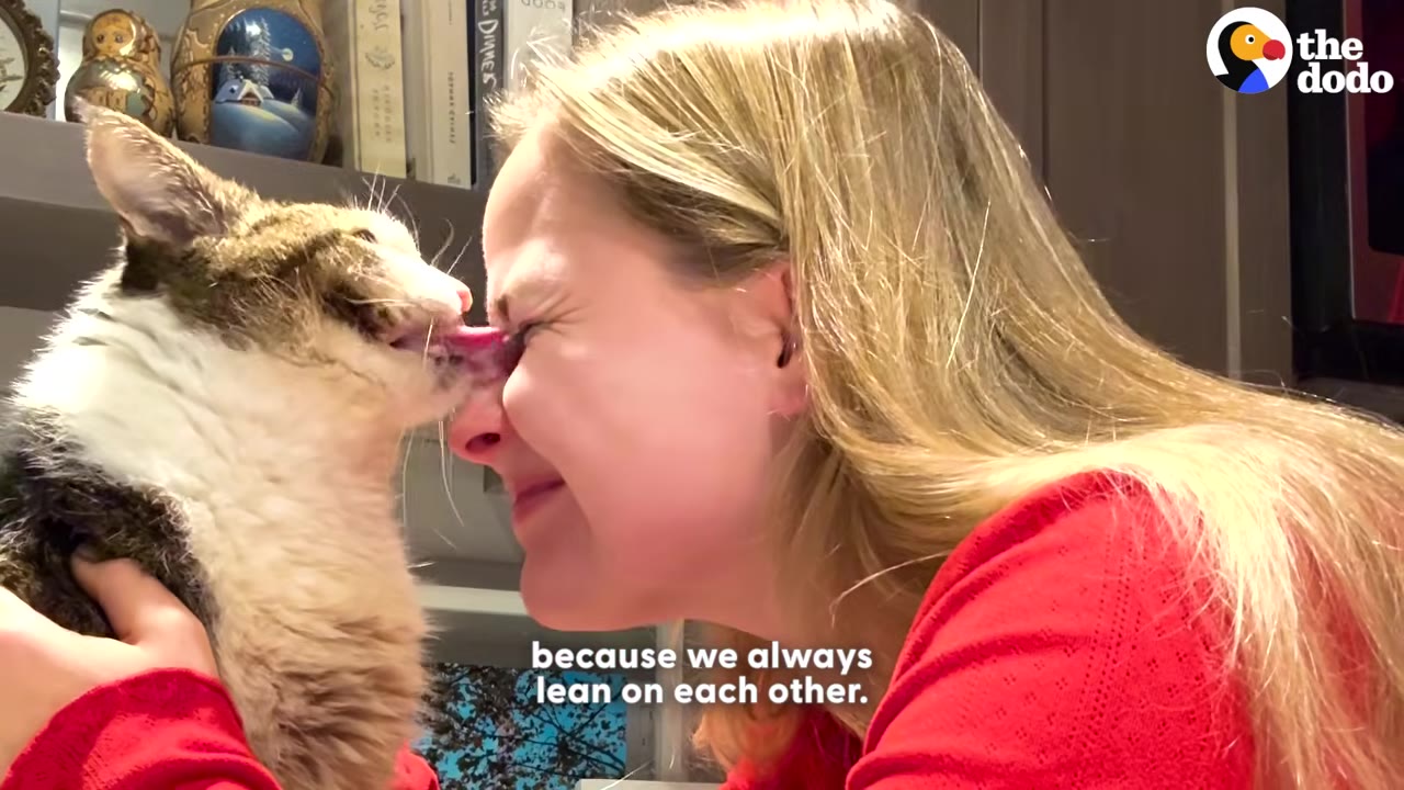 A close-up of a blonde woman in a red shirt holding a brown and white cat close to her face as it licks her nose, with the caption 'because we always lean on each other' at the bottom.
