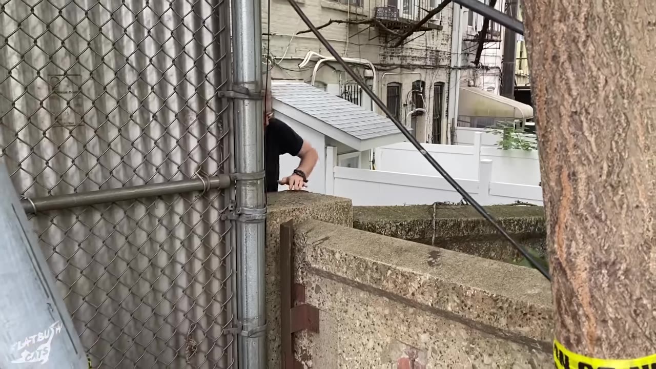 A man in a black t-shirt leans over a concrete wall next to a chain-link fence, reaching down into a hidden area.