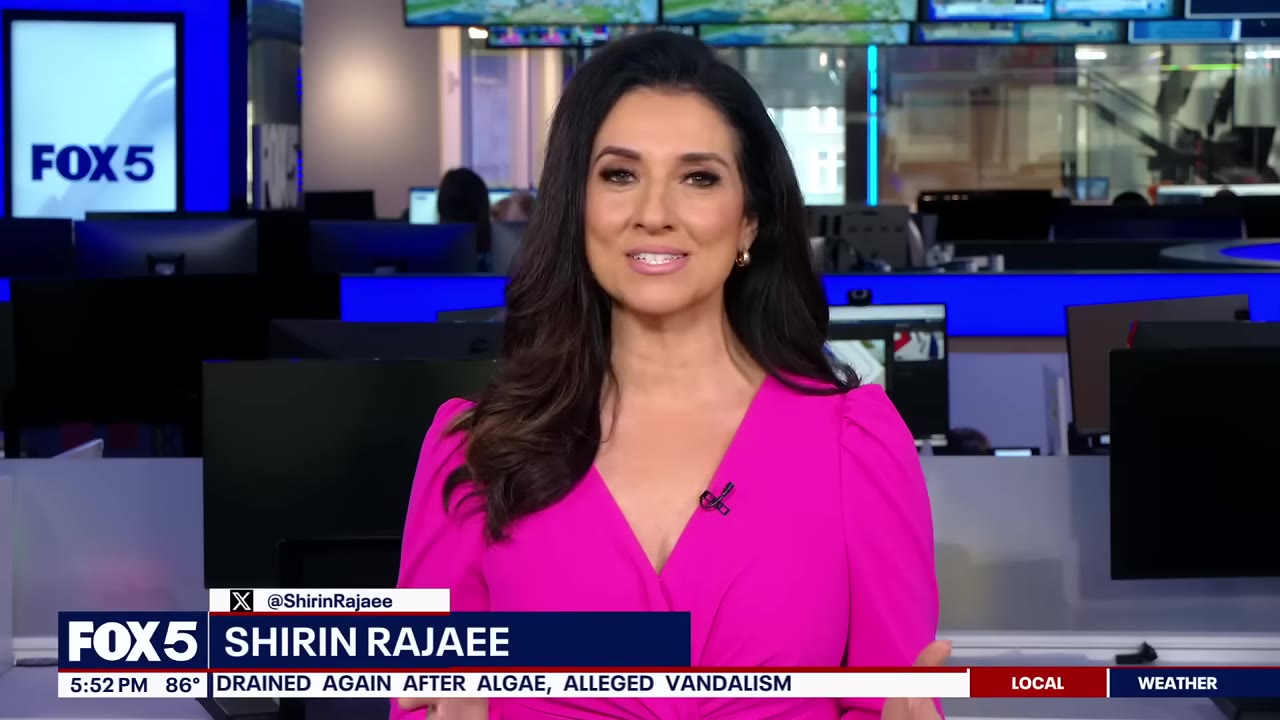 A news anchor with long dark hair wearing a pink dress speaks in a television studio with computer monitors in the background.