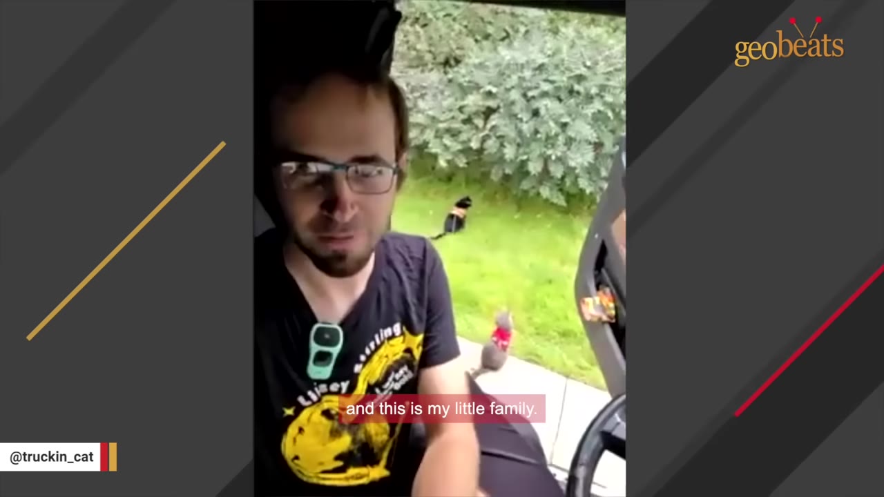 A man wearing glasses and a black graphic t-shirt sits inside a truck cab, looking at the camera, while a cat is visible outside on the grass.