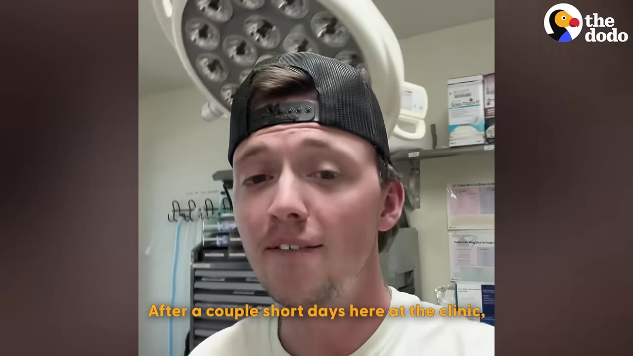 A close-up of a man wearing a black baseball cap backwards, speaking in a clinic room with medical equipment in the background.
