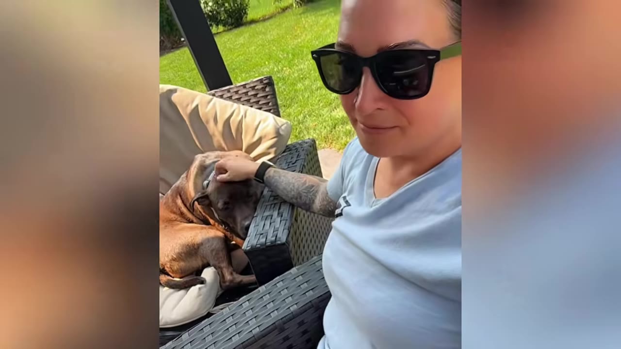A woman wearing sunglasses and a light blue t-shirt sits outdoors, gently petting a brown dog resting on a cushioned patio chair.