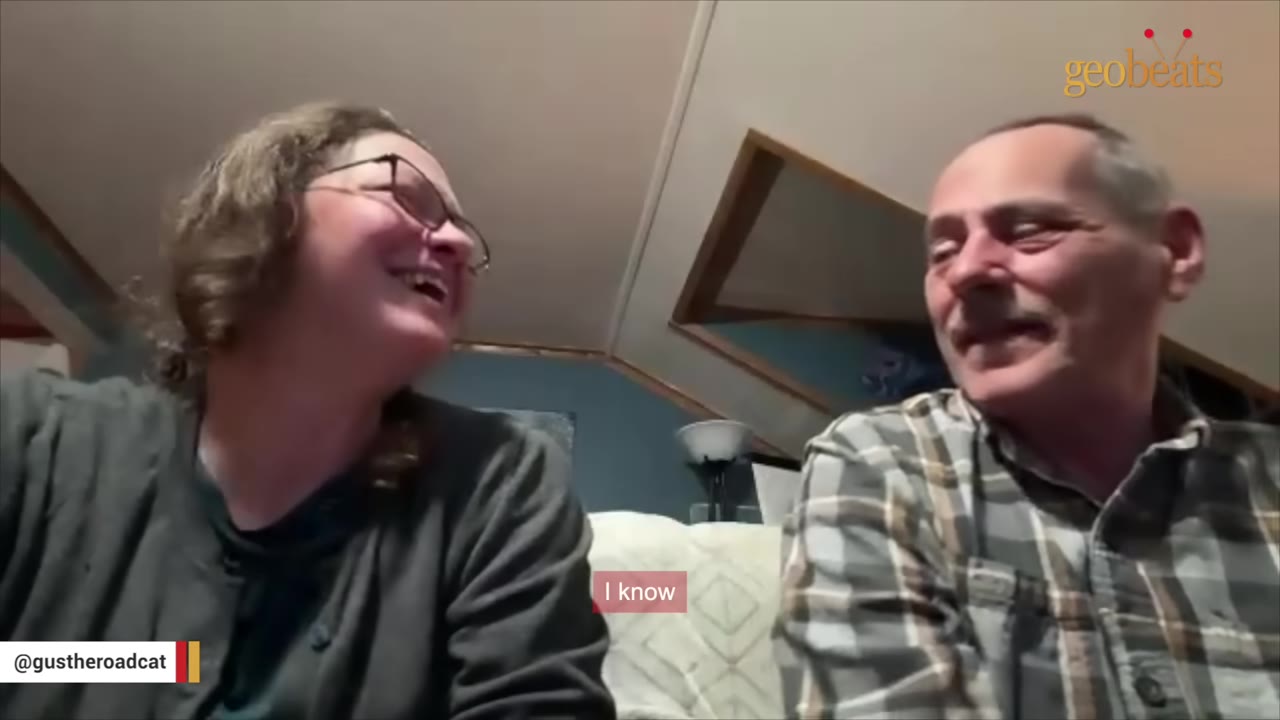A smiling woman with glasses and a man in a plaid shirt look at each other warmly inside a home.