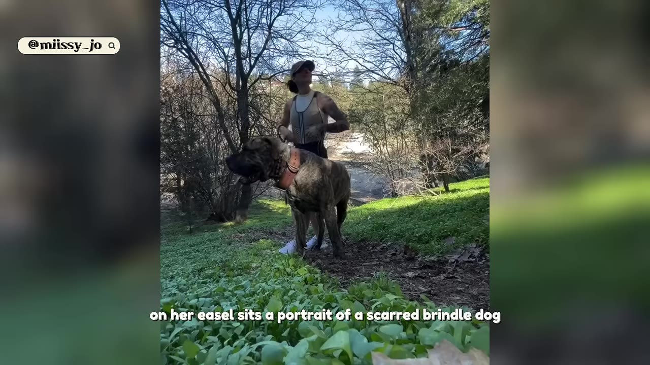 A woman wearing a cap and athletic wear stands in a wooded grassy area next to a large brindle dog on a leash.