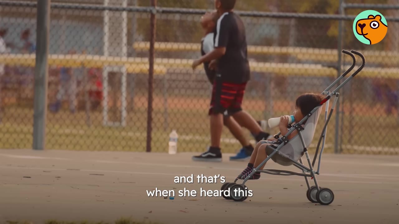 A young child sits in a stroller drinking from a bottle on a paved area near a chain-link fence, with other children playing in the background.