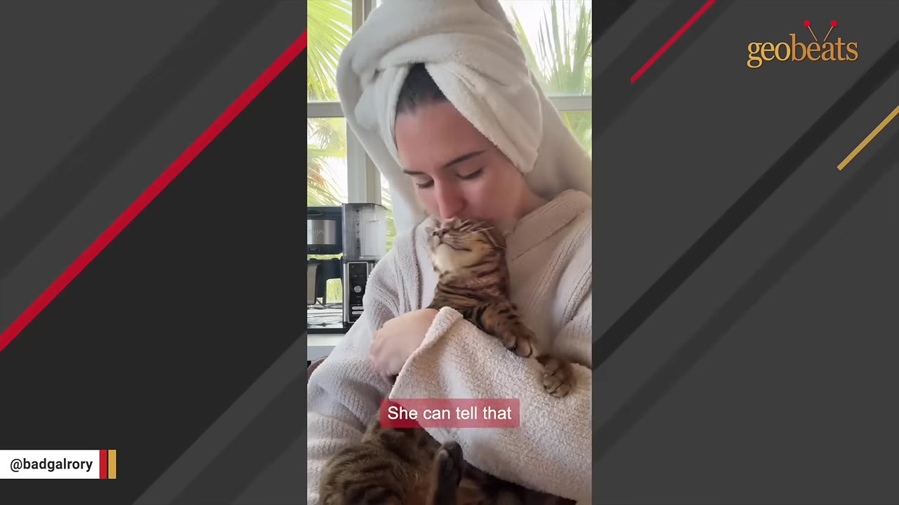 A woman in a white bathrobe and hair towel cradles and kisses a brown tabby cat who has its eyes closed in contentment.