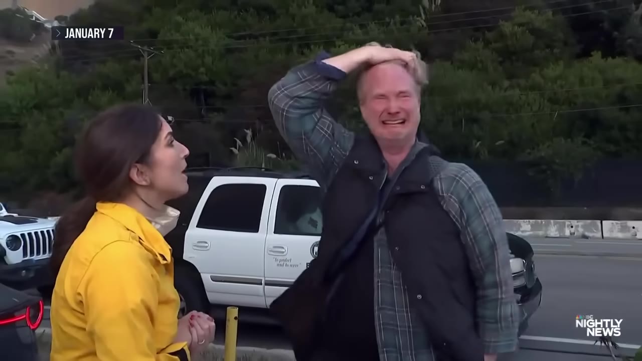 A man crying with his hand on his head in distress next to a reporter in a yellow jacket, with a police vehicle in the background.