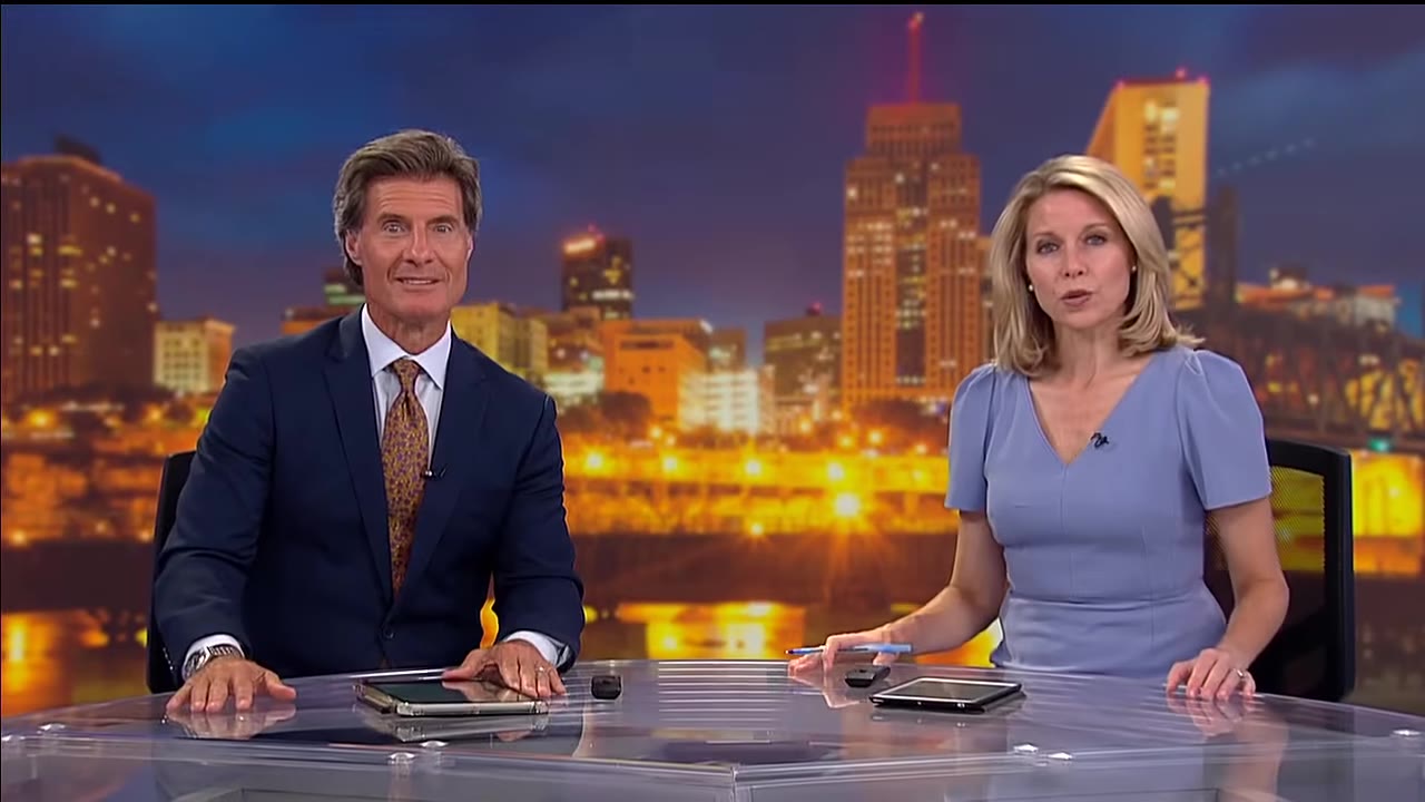 A male and female news anchor sit at a glass desk in front of a city skyline backdrop at night.