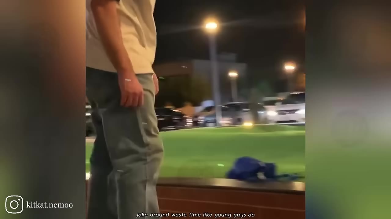 A young man in a light-colored t-shirt and green pants standing outdoors at night near a park or street.