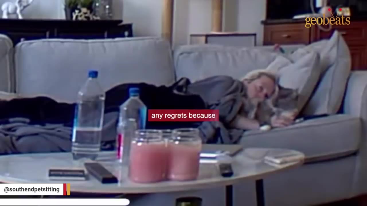 A woman lies on a grey sofa cuddling with a cat, with a coffee table in the foreground holding water bottles and jars.