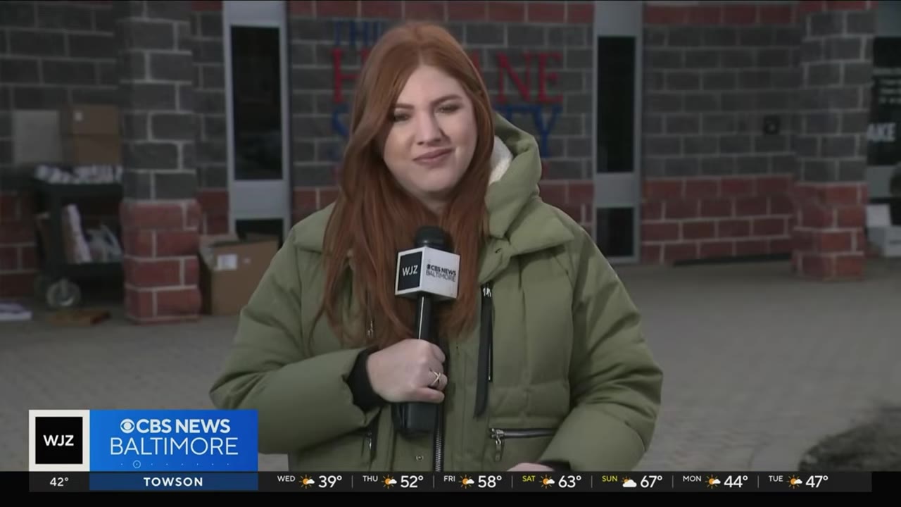 A female news reporter with red hair wearing a green winter coat holds a WJZ CBS News Baltimore microphone while standing outside.