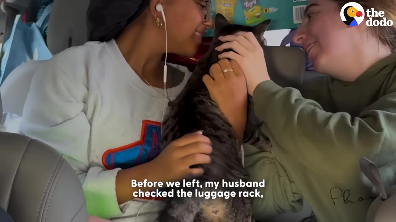 Two young women inside a car smiling and petting a tabby cat they are holding.