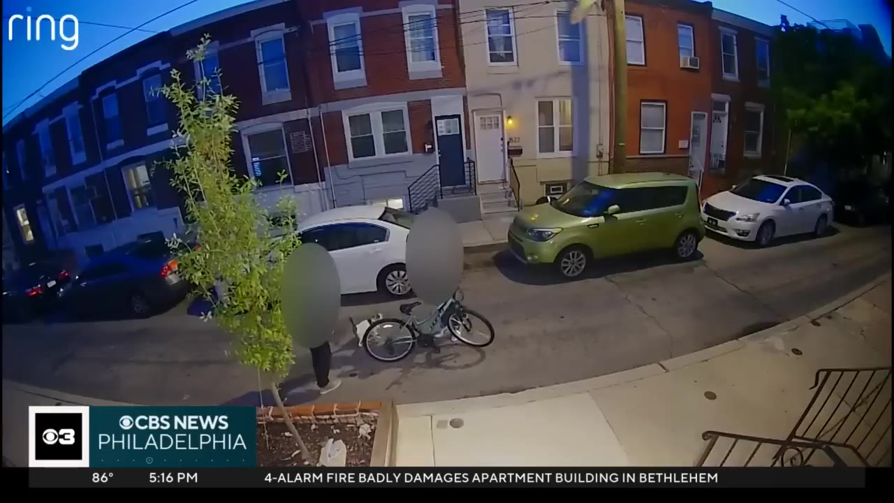 A wide-angle Ring security camera frame showing a residential street with row houses, parked cars, and two individuals with blurred faces standing near a bicycle.