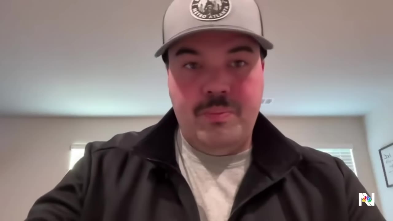 A close-up portrait of a man with a mustache wearing a grey cap and a black jacket, looking directly forward.