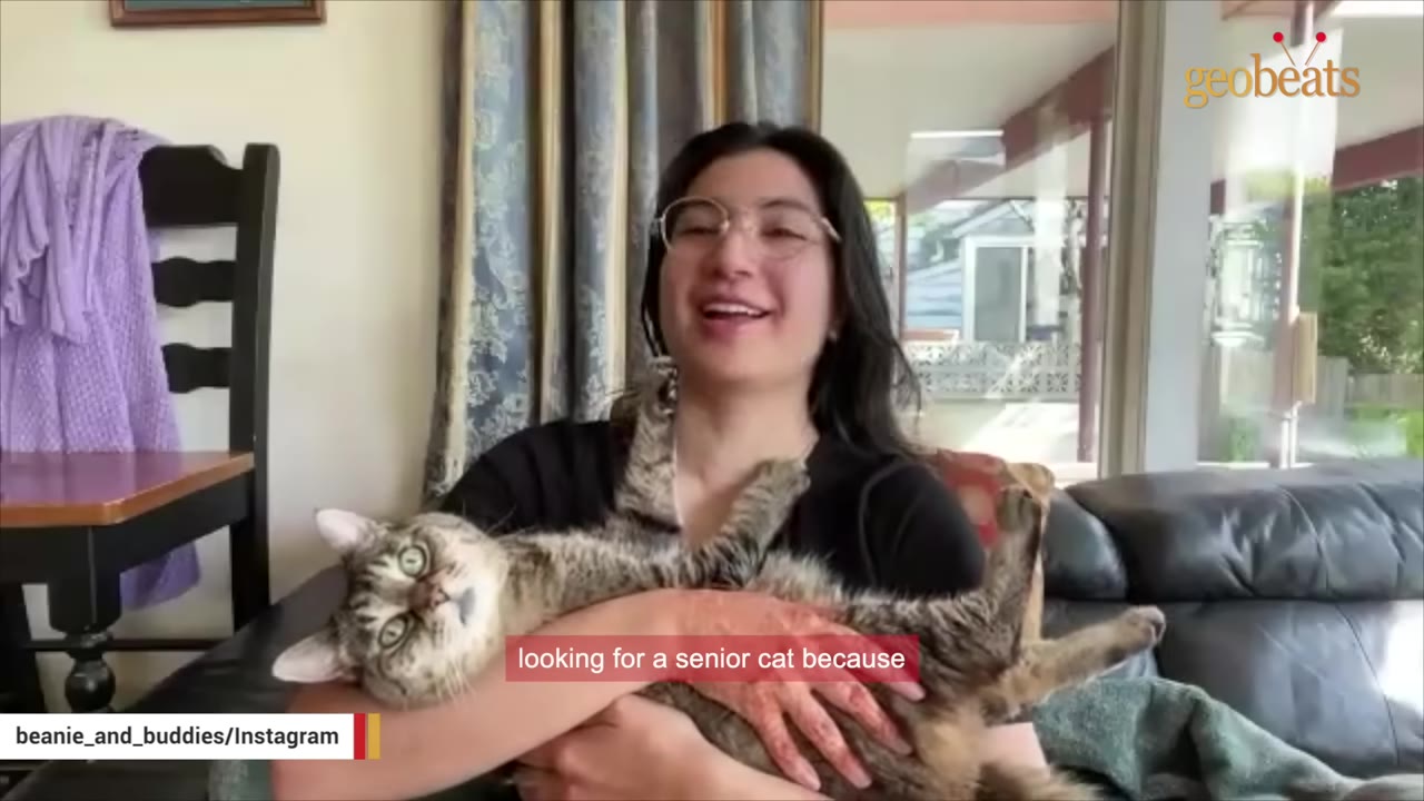 A smiling woman with glasses holding a tabby cat in her arms while sitting on a couch.