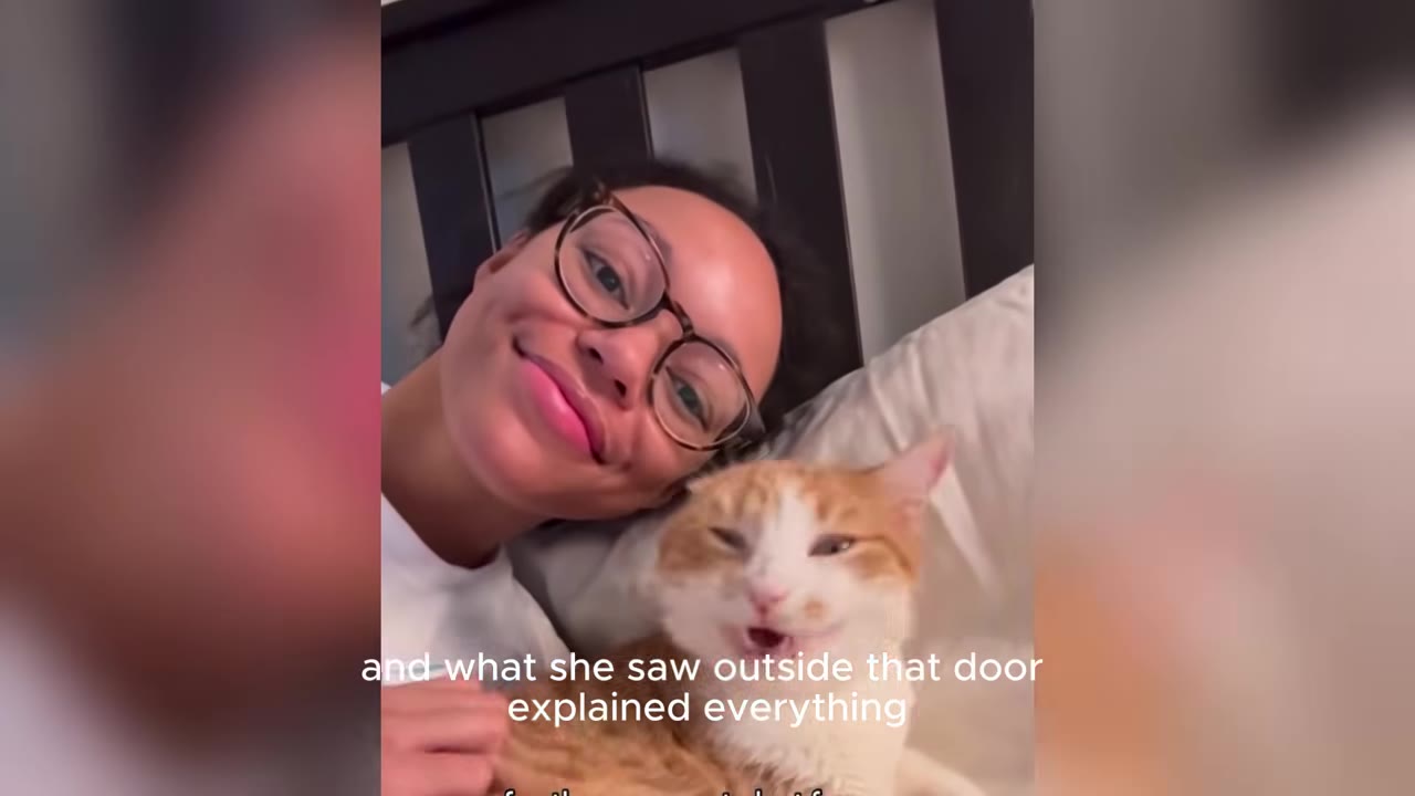A smiling woman wearing glasses lies in bed next to an orange and white cat.