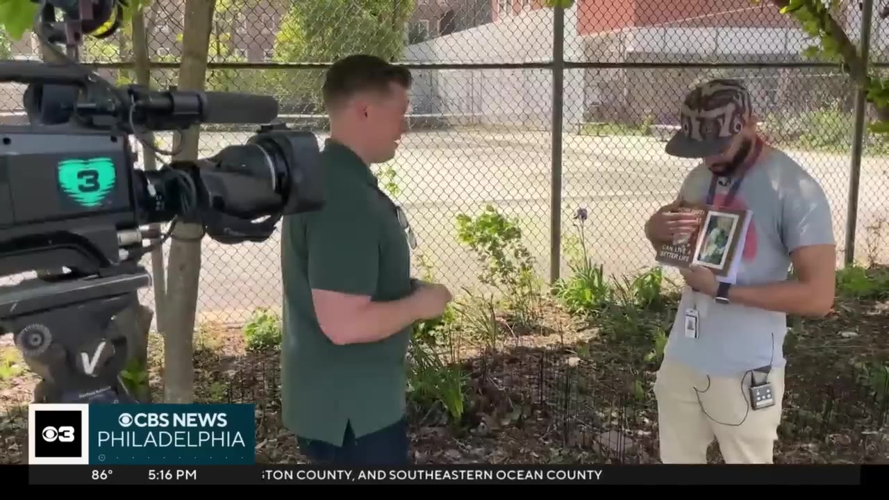 A news reporter in a green shirt interviews a man wearing a cap who is holding a framed picture of a cat, with a television camera visible in the foreground.