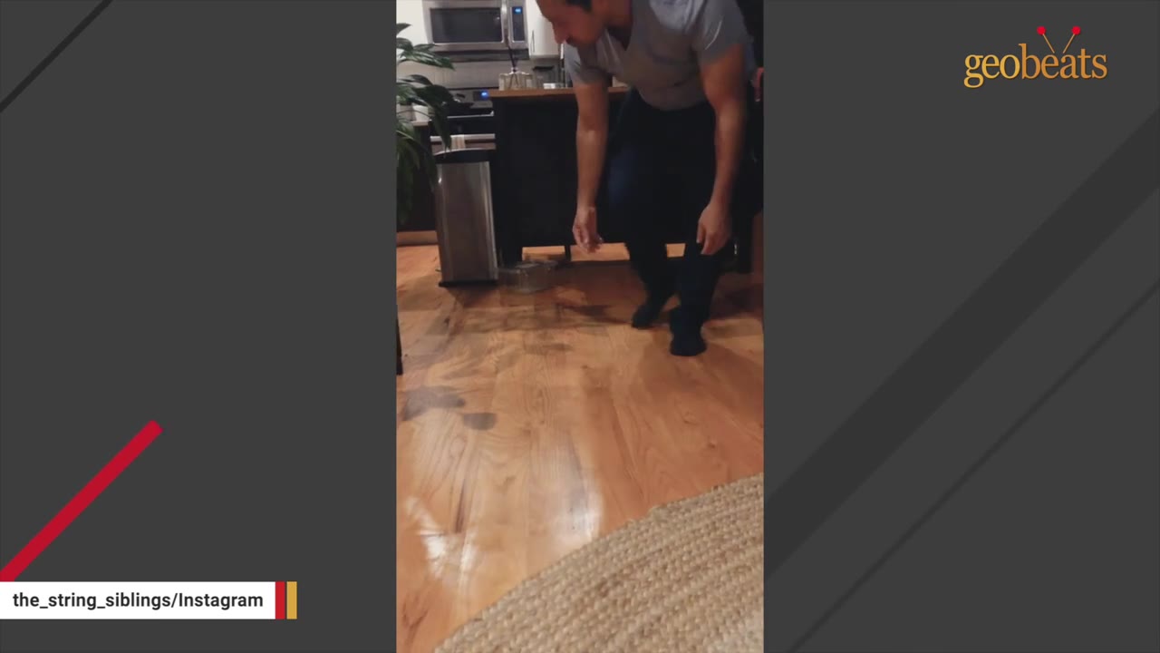 A man in a grey t-shirt and dark pants stands in a kitchen, looking down hesitantly at the wooden floor.
