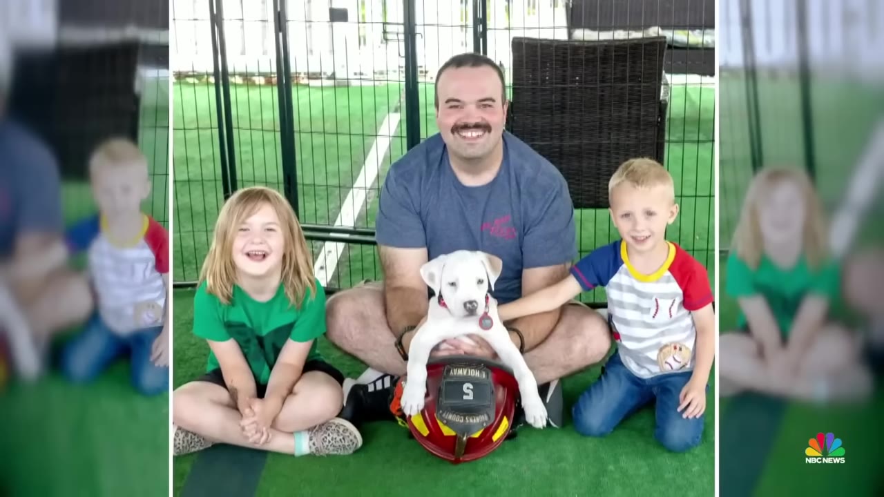 A smiling man sits cross-legged on green turf with two young children, holding a white puppy that is resting its paws on a red firefighter helmet.