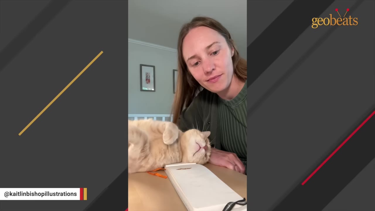 An orange cat lies relaxed on its back on a desk next to an open sketchbook, while a woman looks down at him with a smile.