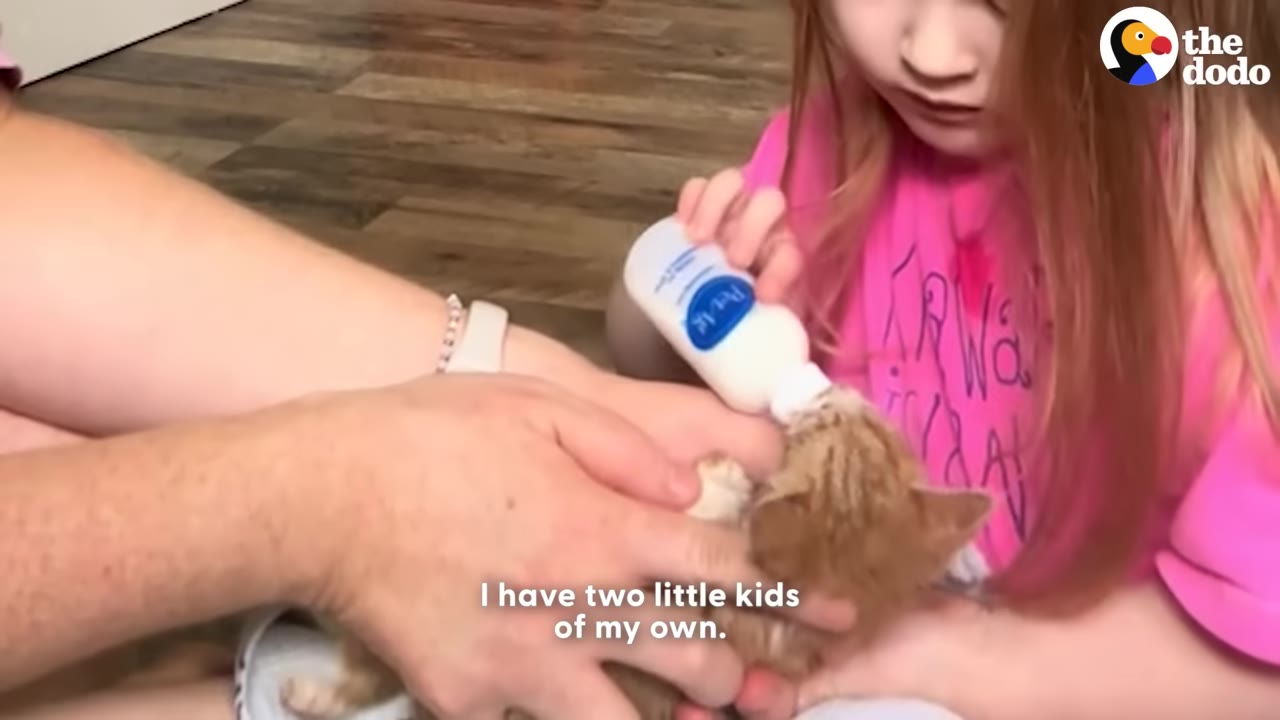 A young girl with long reddish hair wearing a pink shirt holds a small bottle to feed a tiny orange kitten, while an adult's hands help support the kitten.